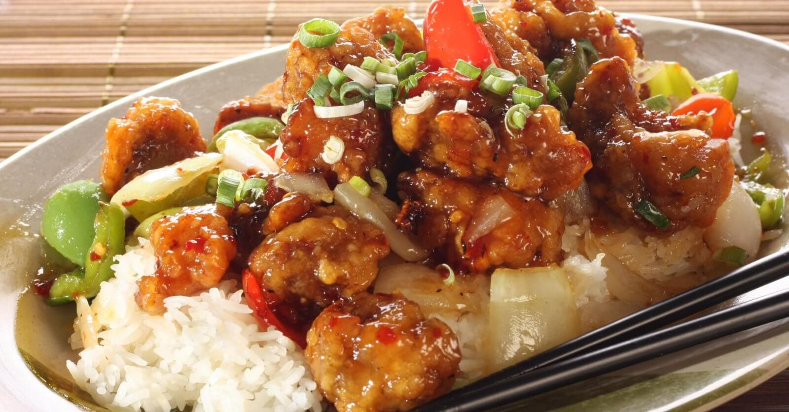 A plate filled with sweet and sour chicken served over white rice is garnished with chopped green onions. The dish includes chunks of chicken coated in a glossy sauce with red and green bell peppers and onions. Black chopsticks rest on the side of the plate, which sits on a wooden surface at Lucky Chinatown.