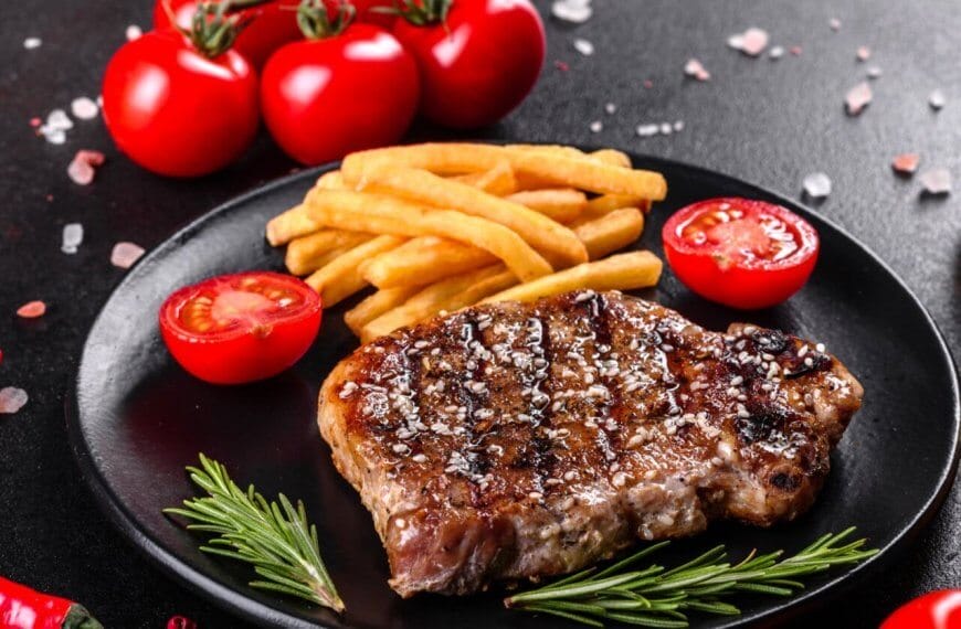 A grilled steak garnished with coarse salt and black pepper sits on a black plate alongside golden French fries and cherry tomato halves, promising a Sulit Dining Experience. Fresh rosemary sprigs lay next to the steak. The background features whole cherry tomatoes, a red chili pepper, sea salt, and a bottle of olive oil.