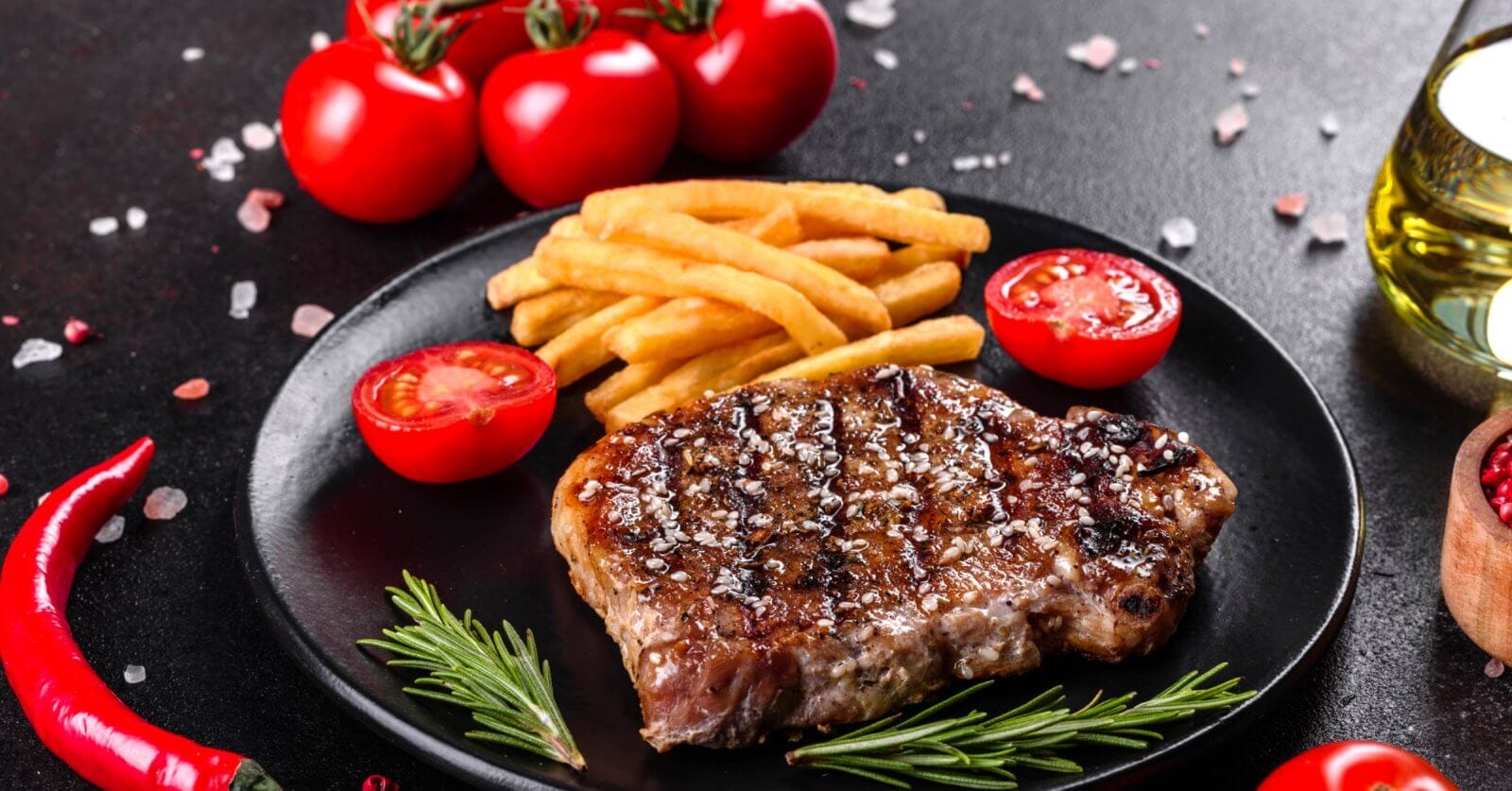 A grilled steak garnished with coarse salt and black pepper sits on a black plate alongside golden French fries and cherry tomato halves, promising a Sulit Dining Experience. Fresh rosemary sprigs lay next to the steak. The background features whole cherry tomatoes, a red chili pepper, sea salt, and a bottle of olive oil.