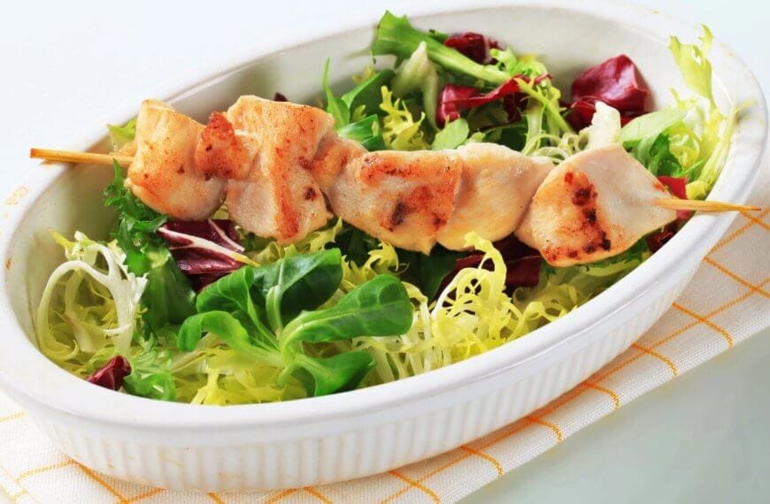 A white oval dish contains a fresh salad with green and purple lettuce, topped with a skewer of grilled chicken pieces. The dish, representing the best of sulit dining in Baguio, is placed on a folded, white napkin with yellow lines. The tender, slightly charred chicken contrasts with the vibrant greens of the salad.