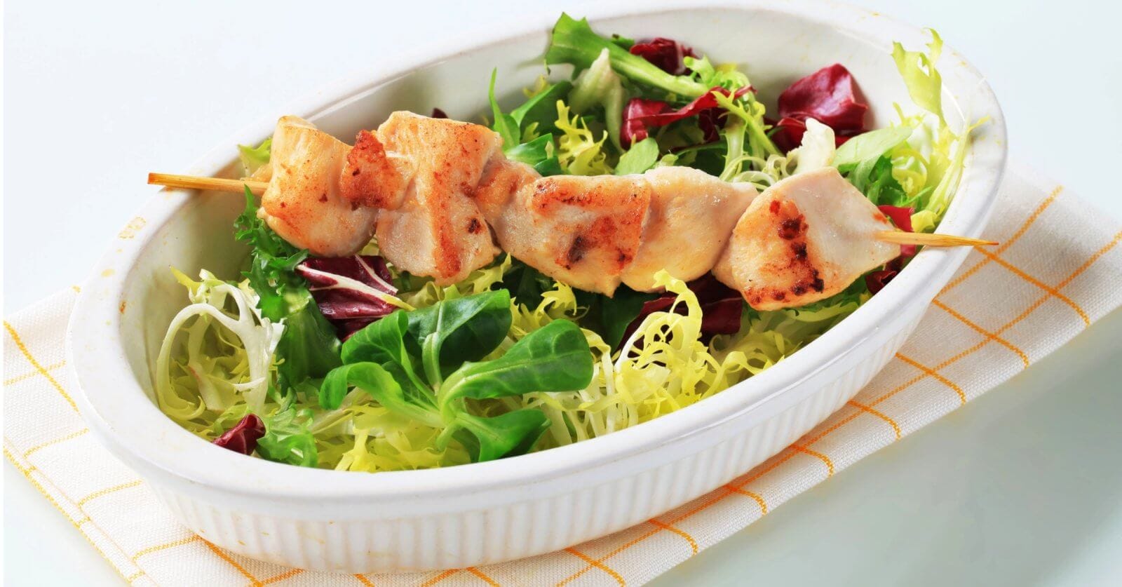 A white oval dish contains a fresh salad with green and purple lettuce, topped with a skewer of grilled chicken pieces. The dish, representing the best of sulit dining in Baguio, is placed on a folded, white napkin with yellow lines. The tender, slightly charred chicken contrasts with the vibrant greens of the salad.