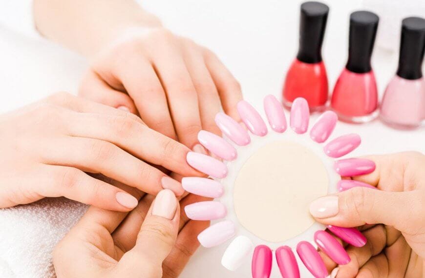 At one of the best nail lounges in Metro Manila, a person indulges in a relaxing manicure. Another holds a nail color palette of various pink and white shades against their nails. In the background, several bottles of red, pink, and beige nail polish sit on a pristine white surface, completing the spa-like ambiance.