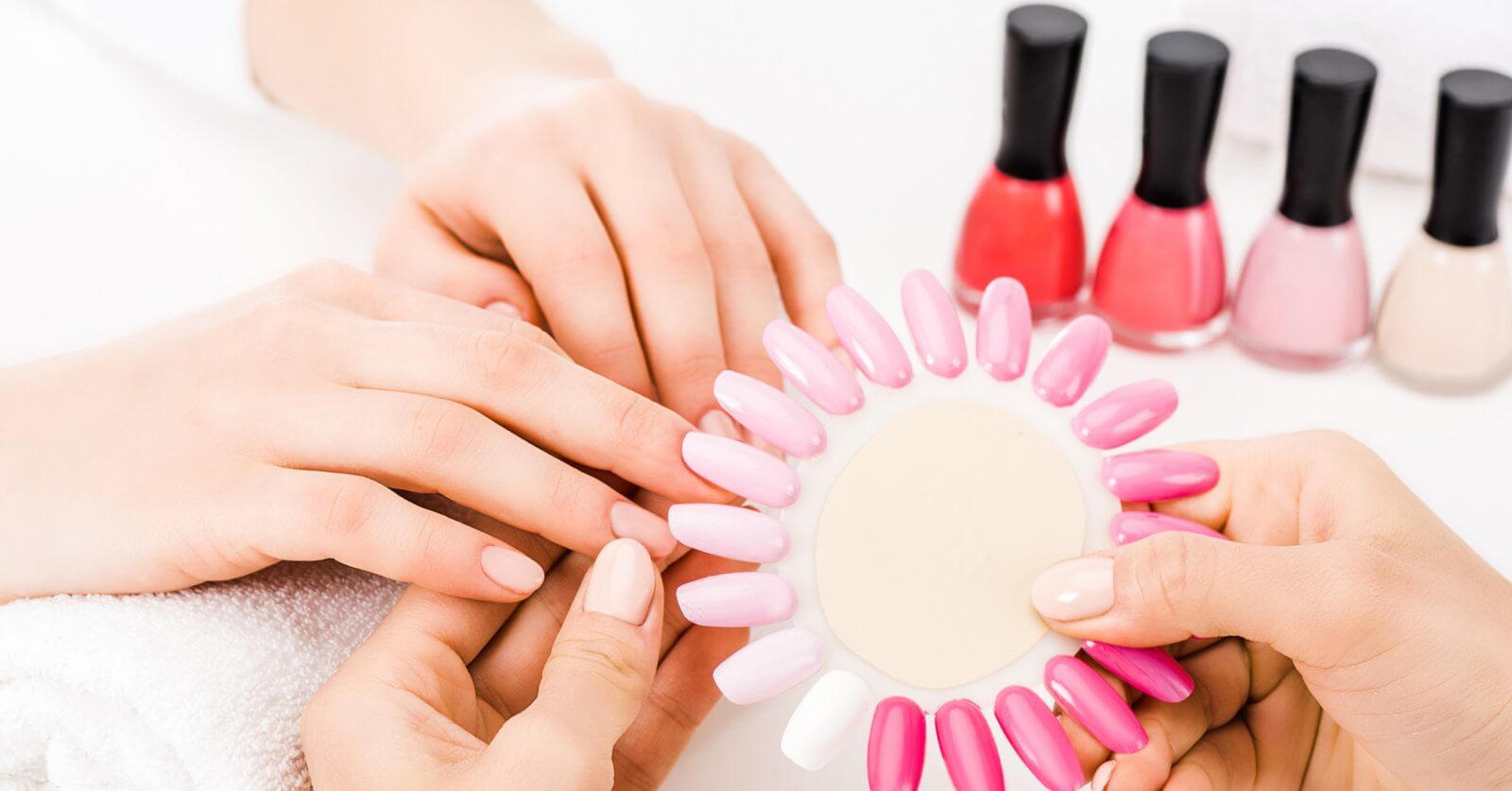 At one of the best nail lounges in Metro Manila, a person indulges in a relaxing manicure. Another holds a nail color palette of various pink and white shades against their nails. In the background, several bottles of red, pink, and beige nail polish sit on a pristine white surface, completing the spa-like ambiance.