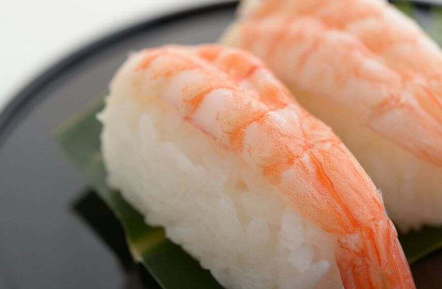 Close-up of two pieces of shrimp nigiri sushi on a black plate. Each sushi piece consists of a cooked shrimp placed atop a small mound of white rice, both resting on a green leaf. This tempting treat is part of the Food Lover's Guide at City of Dreams Manila. The background is blurred with a white backdrop.