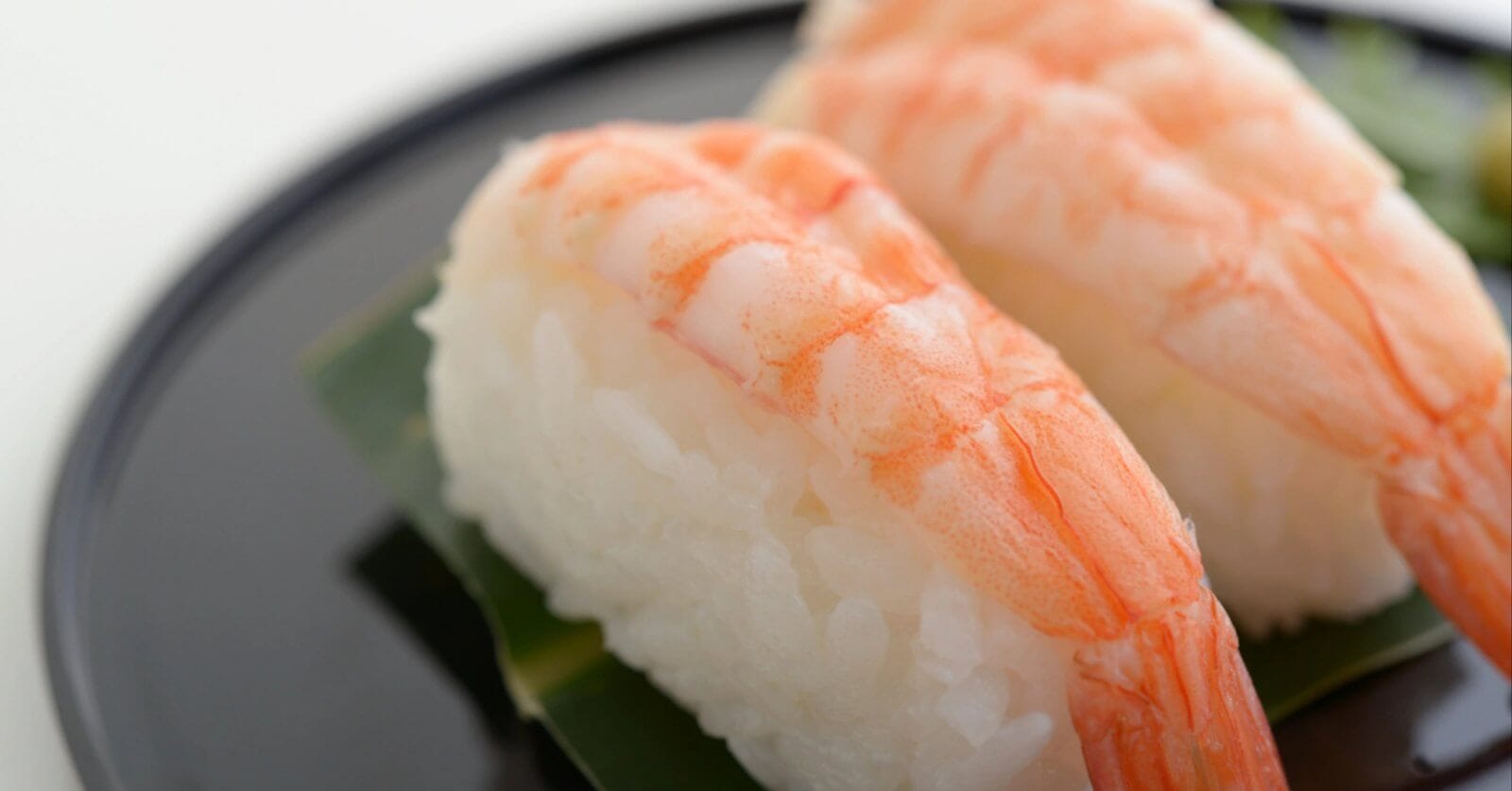 Close-up of two pieces of shrimp nigiri sushi on a black plate. Each sushi piece consists of a cooked shrimp placed atop a small mound of white rice, both resting on a green leaf. This tempting treat is part of the Food Lover's Guide at City of Dreams Manila. The background is blurred with a white backdrop.