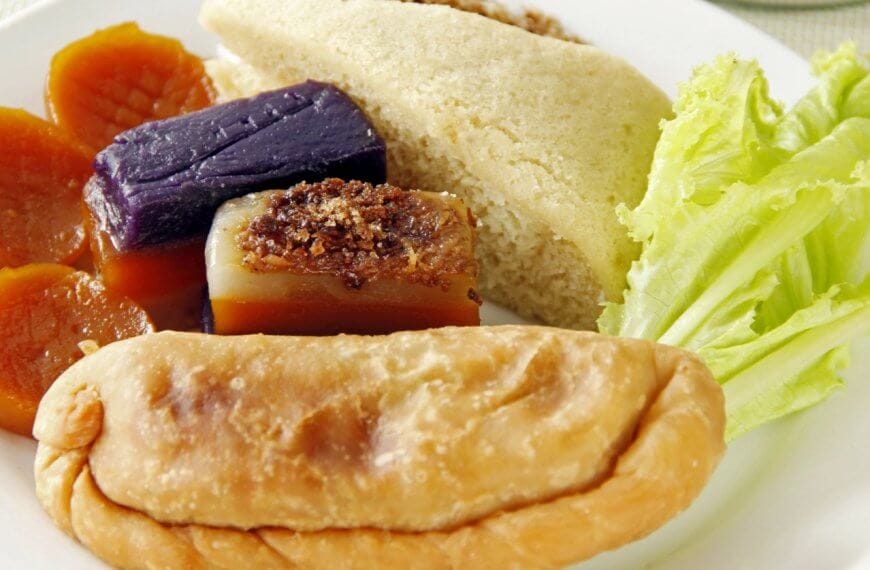 A plate with a variety of Filipino snacks, including a fried empanada in the foreground, slices of puto cake, sapin-sapin in purple and white, slices of orange kamote (sweet potato), and a piece of fresh lettuce on the side, all arranged on a white plate. Perfect for enjoying at one of Quezon City's best restaurants near Robinsons Magnolia.