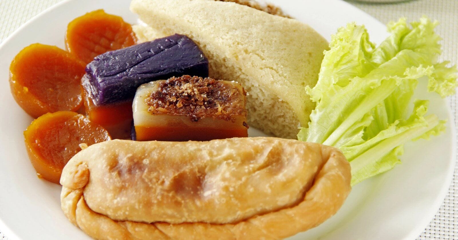 A plate with a variety of Filipino snacks, including a fried empanada in the foreground, slices of puto cake, sapin-sapin in purple and white, slices of orange kamote (sweet potato), and a piece of fresh lettuce on the side, all arranged on a white plate. Perfect for enjoying at one of Quezon City's best restaurants near Robinsons Magnolia.