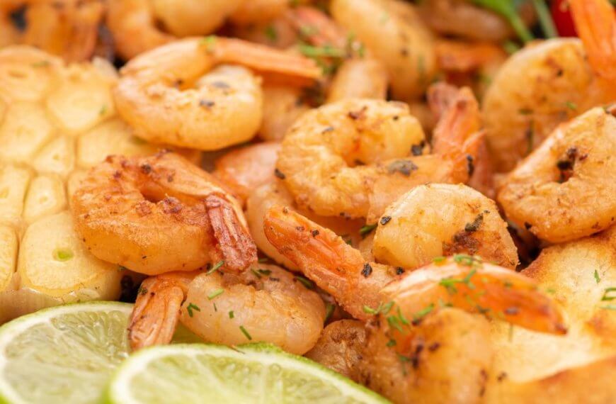 Close-up image of a plate of grilled shrimp garnished with chopped herbs. The shrimp are accompanied by toasted bread slices, grilled garlic, lime slices, and colorful vegetables like cherry tomatoes and bell peppers. This appetizing and well-seasoned dish offers a sulit experience reminiscent of top seafood restaurants in Metro Manila.
