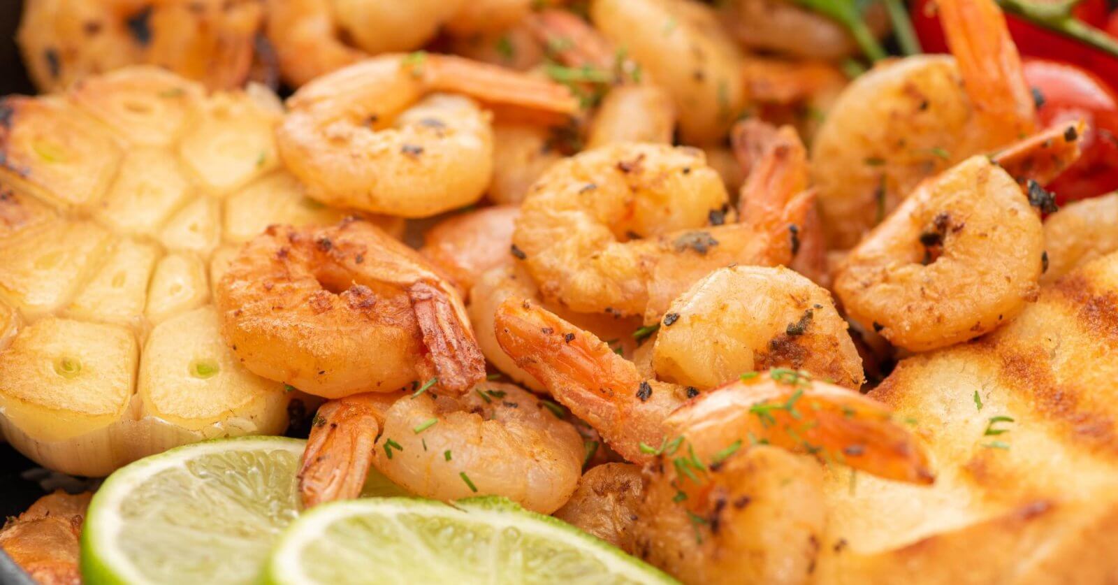 Close-up image of a plate of grilled shrimp garnished with chopped herbs. The shrimp are accompanied by toasted bread slices, grilled garlic, lime slices, and colorful vegetables like cherry tomatoes and bell peppers. This appetizing and well-seasoned dish offers a sulit experience reminiscent of top seafood restaurants in Metro Manila.