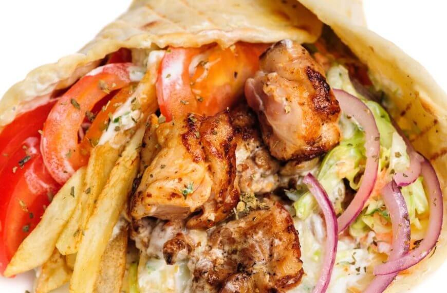 A close-up image of a gyro wrap featuring grilled pieces of seasoned meat, fresh tomato slices, red onion, lettuce, and a generous amount of fries. The ingredients are wrapped in a soft pita bread and drizzled with a creamy white sauce. This dish is one of the best places to enjoy shawarma in Metro Manila.