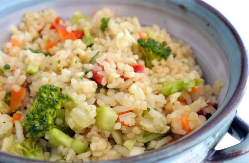 A ceramic bowl filled with a colorful vegetable rice dish, including broccoli florets, diced red bell peppers, and finely chopped carrots. The rice appears fluffy and well-cooked, with visible seasoning interspersed. Evoking the vibe of Katipunan's 63 Best Restaurants at UP Town Center, the bowl has rustic blue and gray tones.