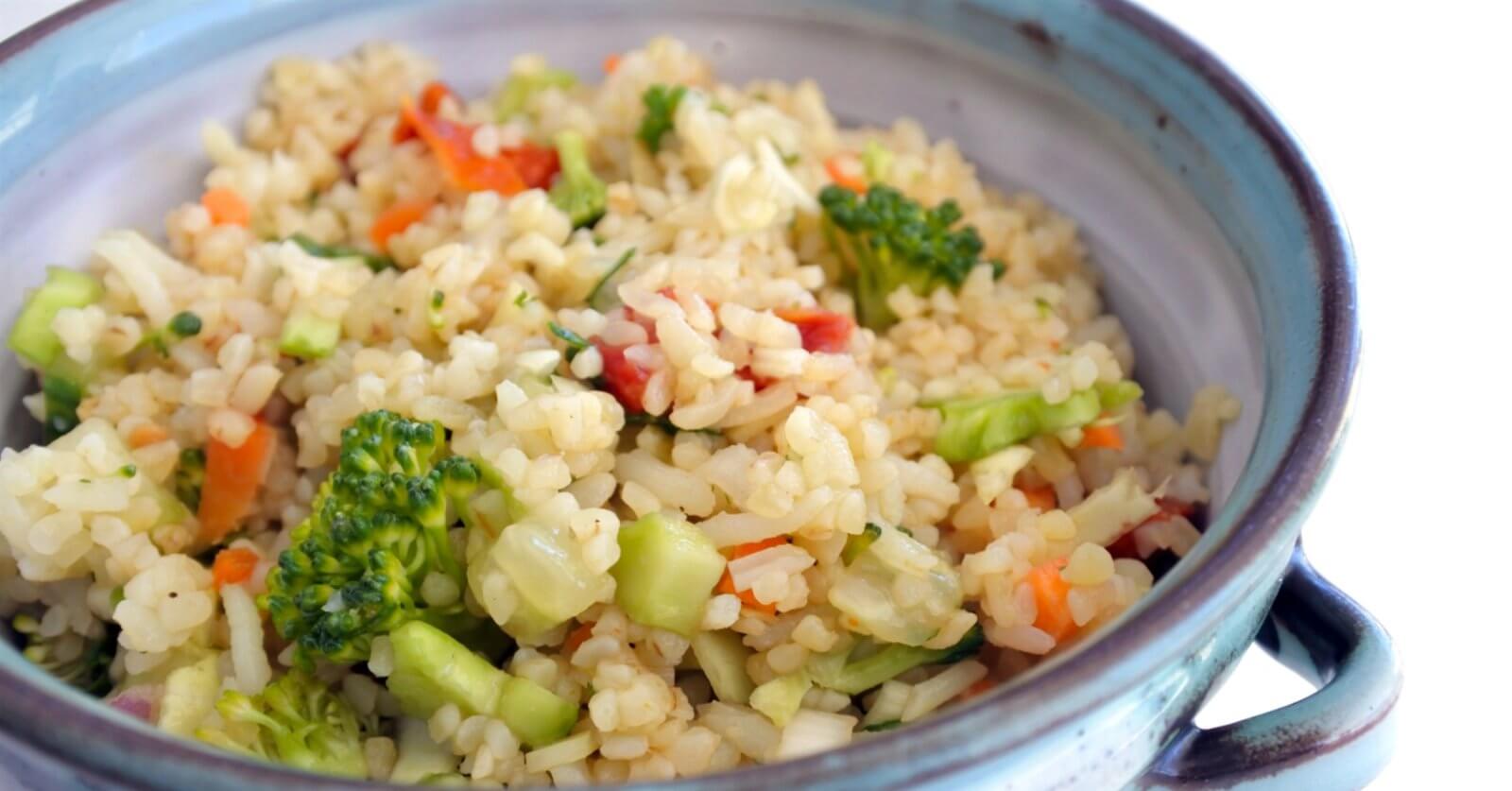 A ceramic bowl filled with a colorful vegetable rice dish, including broccoli florets, diced red bell peppers, and finely chopped carrots. The rice appears fluffy and well-cooked, with visible seasoning interspersed. Evoking the vibe of Katipunan's 63 Best Restaurants at UP Town Center, the bowl has rustic blue and gray tones.