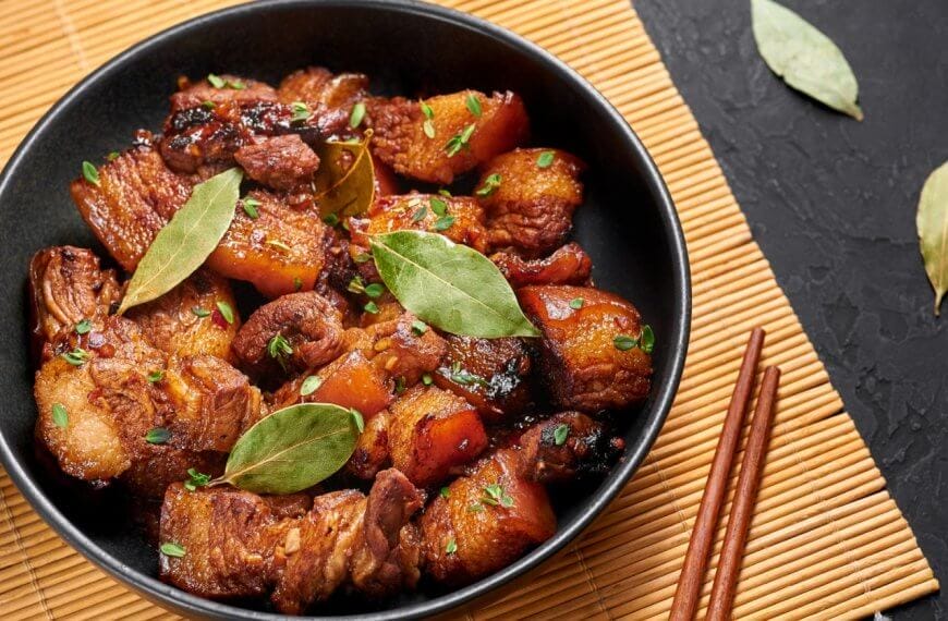 A black bowl filled with caramelized pork belly garnished with green herbs and bay leaves sits on a bamboo mat. At Uptown Mall, BGC, this dish is among the best restaurants' highlights. Two wooden chopsticks lie beside the bowl on a dark, textured surface with loose bay leaves scattered nearby.