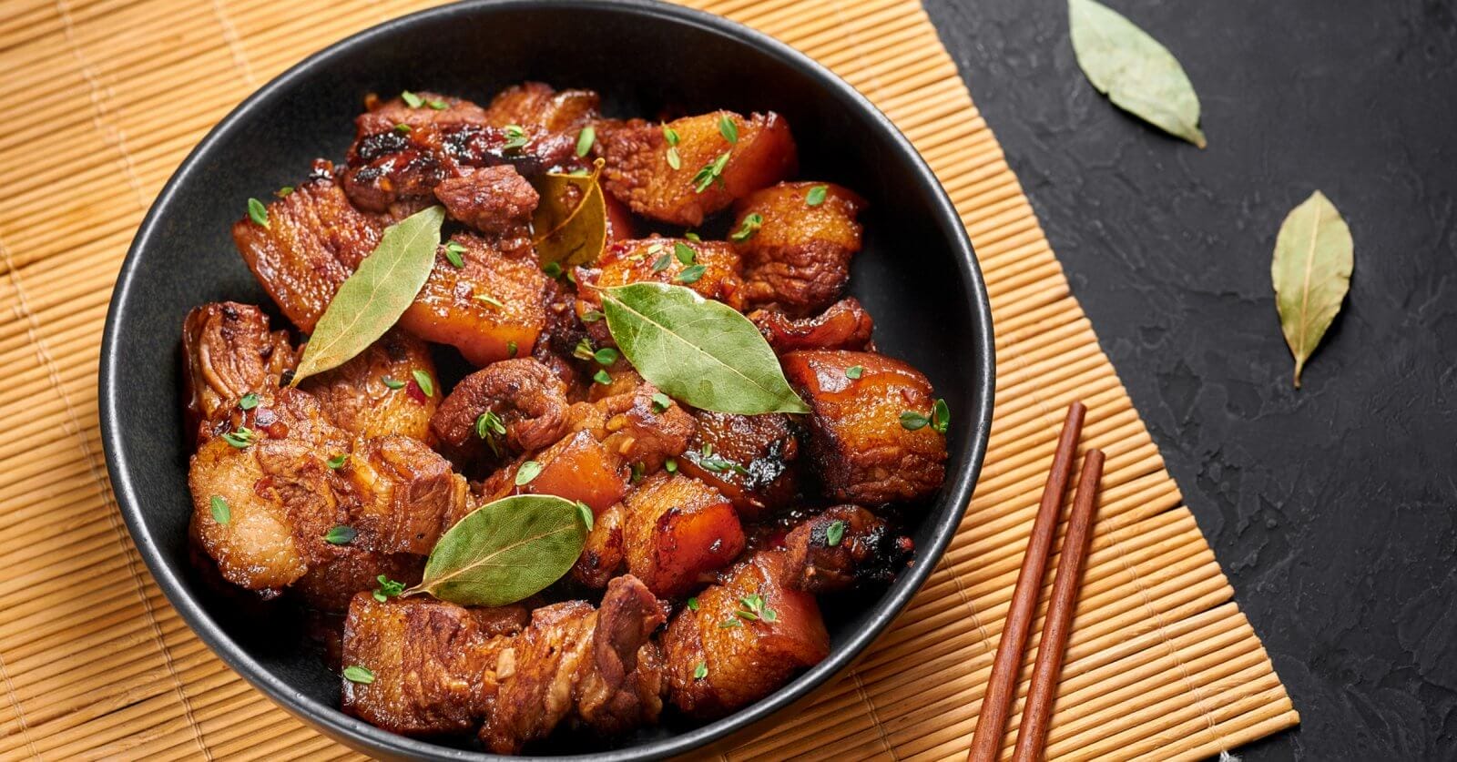 A black bowl filled with caramelized pork belly garnished with green herbs and bay leaves sits on a bamboo mat. At Uptown Mall, BGC, this dish is among the best restaurants' highlights. Two wooden chopsticks lie beside the bowl on a dark, textured surface with loose bay leaves scattered nearby.
