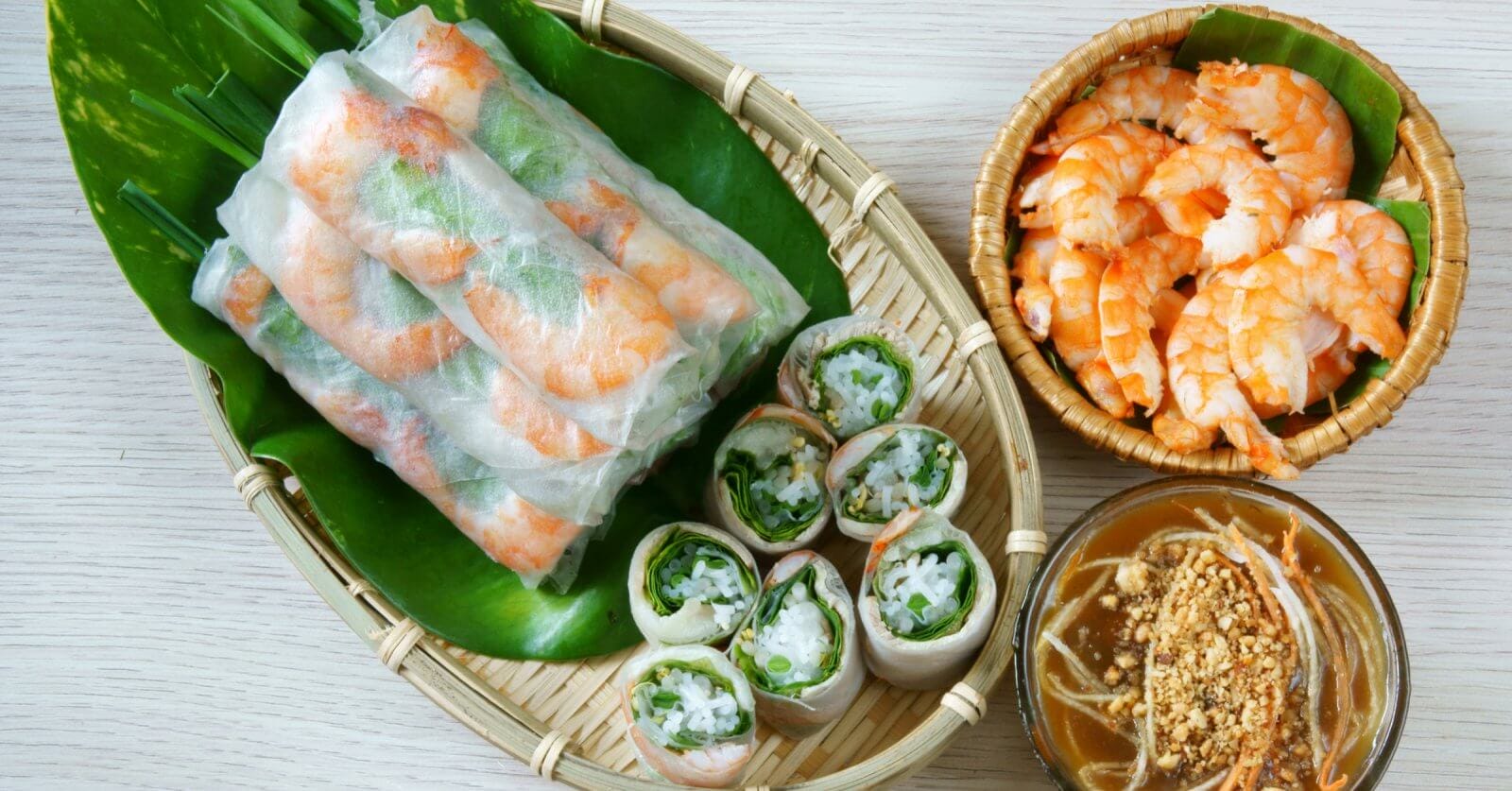 A woven tray holding fresh spring rolls filled with shrimp and vegetables rests atop a green banana leaf. Next to it, a small wicker bowl contains cooked shrimp, and another bowl is filled with brown peanut sauce garnished with crushed peanuts and sliced vegetables—a nod to authentic dining in Metro Manila's Vietnamese restaurants.