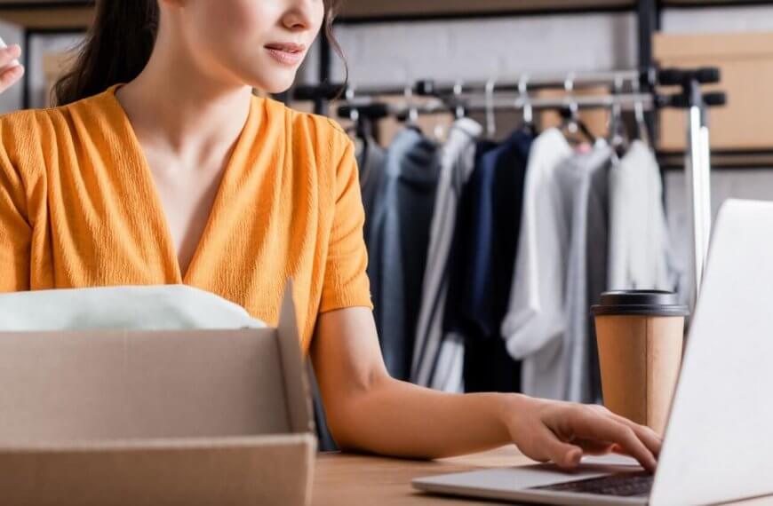 A woman in a yellow dress is sitting at a desk, holding a smartphone and typing on a laptop. In front of her is an open cardboard box, and a cup of coffee sits next to her computer. Behind her, a clothing rack with various garments is visible, suggesting she's running a small online business.