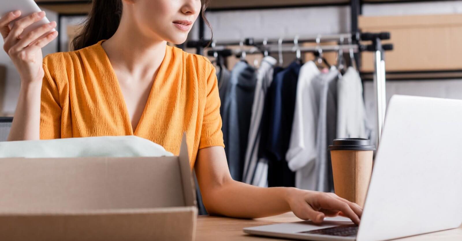 A woman in a yellow dress is sitting at a desk, holding a smartphone and typing on a laptop. In front of her is an open cardboard box, and a cup of coffee sits next to her computer. Behind her, a clothing rack with various garments is visible, suggesting she's running a small online business.