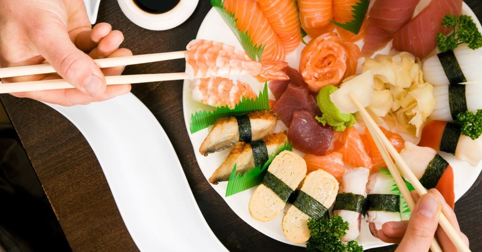 A variety of sushi and sashimi arranged on a white plate, including salmon, tuna, shrimp, and eel nigiri, tamago, and garnishes of wasabi, pickled ginger, and parsley. Two pairs of chopsticks are picking pieces from the platter at one of the restaurants in Ayala Alabang Town Center. A soy sauce dish is nearby on a dark wooden table.