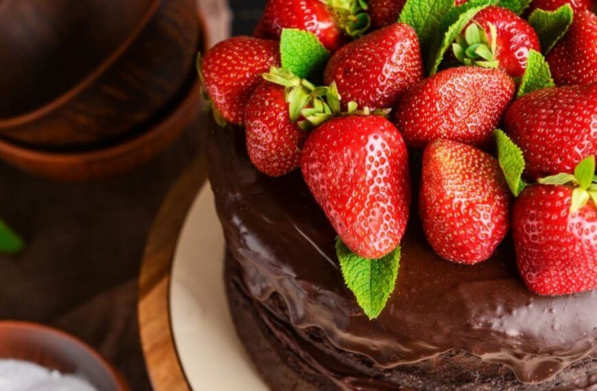 A close-up of a chocolate cake topped with a generous amount of fresh, red strawberries and green mint leaves. The cake has a shiny, smooth chocolate glaze. Brown bowls and green mint leaves are blurred in the background, perfect for those visiting the best restaurants at Ayala Feliz Mall.