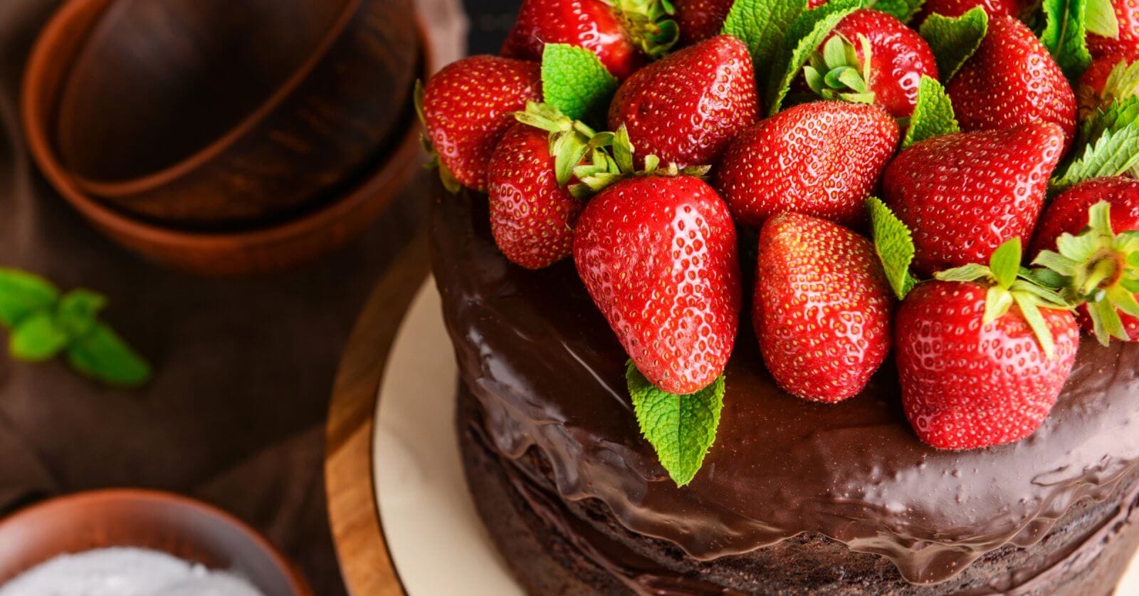 A close-up of a chocolate cake topped with a generous amount of fresh, red strawberries and green mint leaves. The cake has a shiny, smooth chocolate glaze. Brown bowls and green mint leaves are blurred in the background, perfect for those visiting the best restaurants at Ayala Feliz Mall.