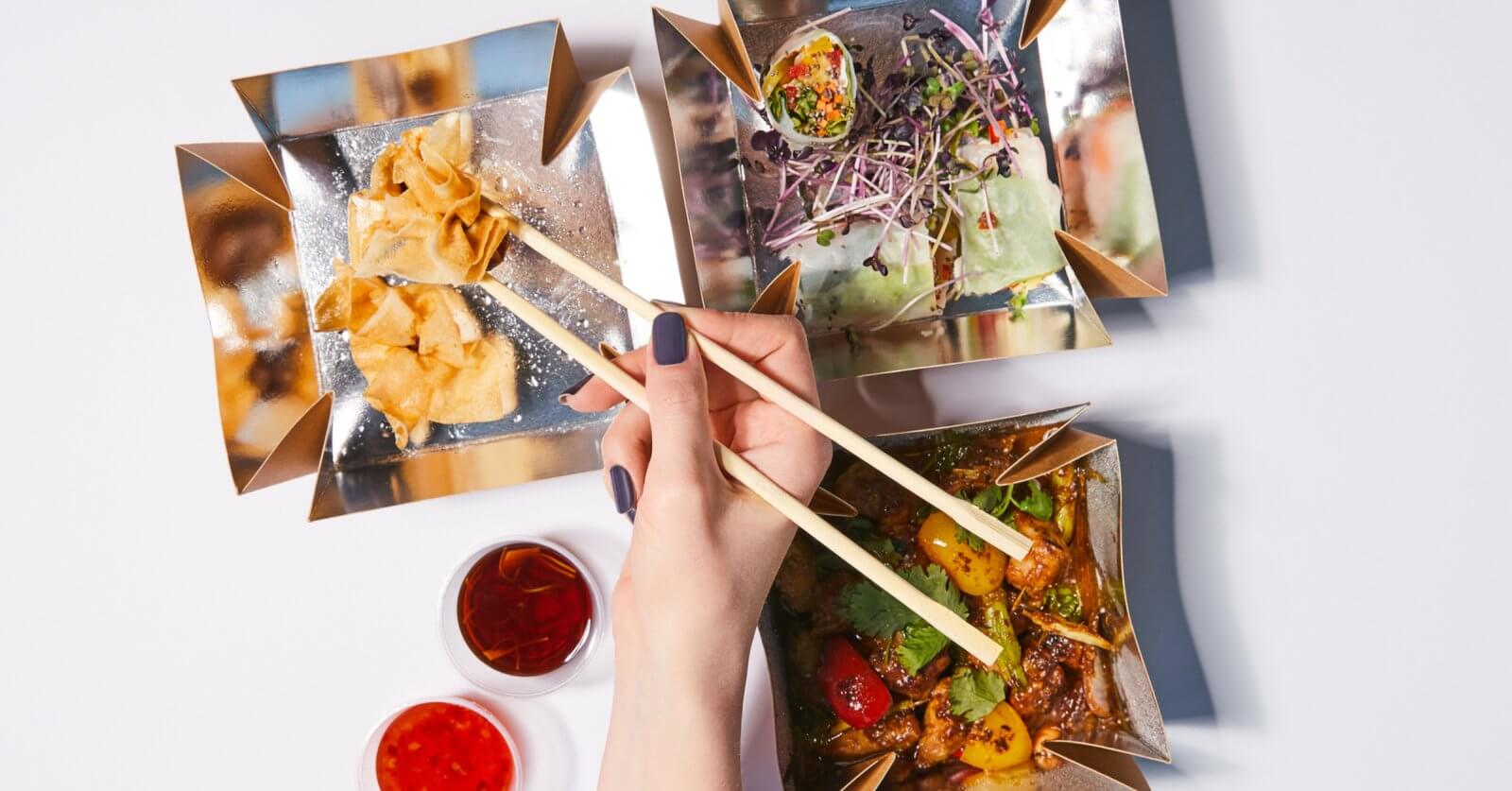 chopsticks to pick up a piece of food from one of three square, gold-colored dishes at Ayala Malls Vermosa. The dishes contain fried wontons, a salad with sprouts, and a cooked vegetable and meat dish. Two small dishes of red dipping sauces are placed nearby on a white surface.