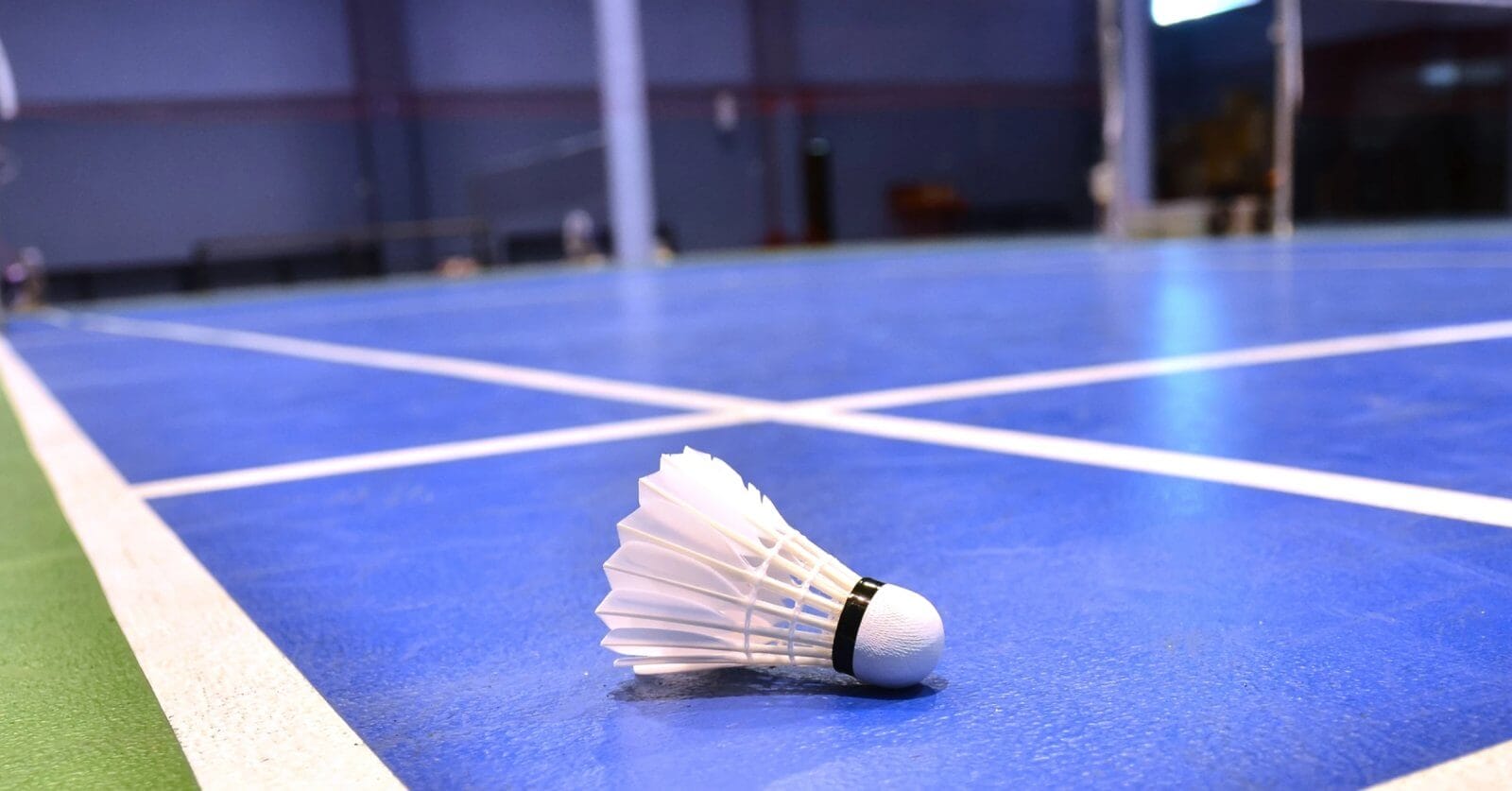 Close-up of a badminton shuttlecock lying on a blue indoor badminton court in Metro Manila. The shuttlecock, with its white feathers and black band, is in the foreground on the court's surface, with the net and other court lines visible in the background. The room has a blurred, spacious interior.