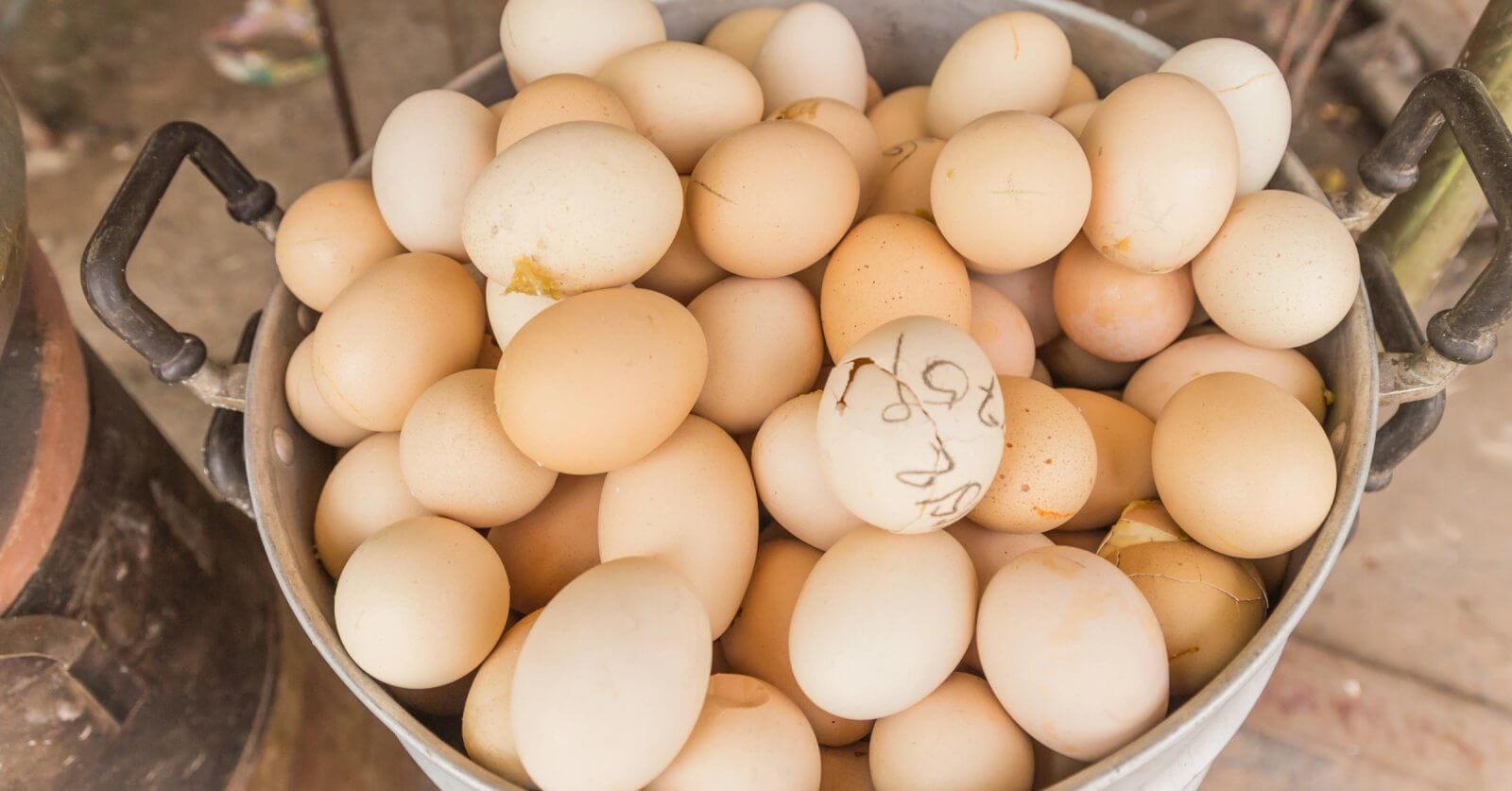 A large metal bucket filled with Balut, some bearing small dirt stains or marks on their shells. The bucket, featuring two handles on either side, rests on a rustic surface, evoking the setting of a farm or market – ideal for Balut suppliers in Metro Manila.