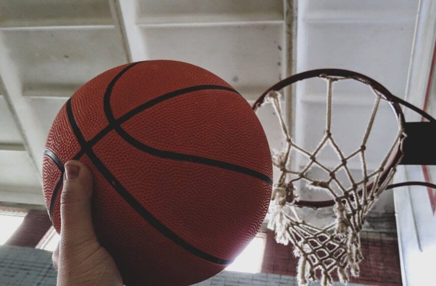 A hand holding an orange basketball is positioned near a hoop on an indoor basketball court available for rent.