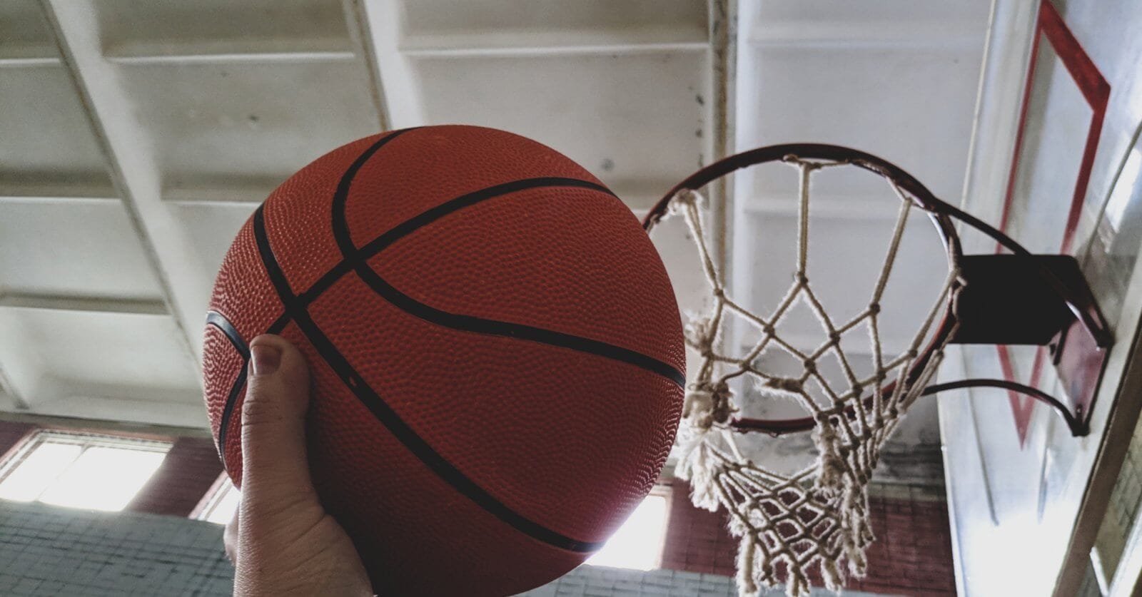 A hand holding an orange basketball is positioned near a hoop on an indoor basketball court available for rent.