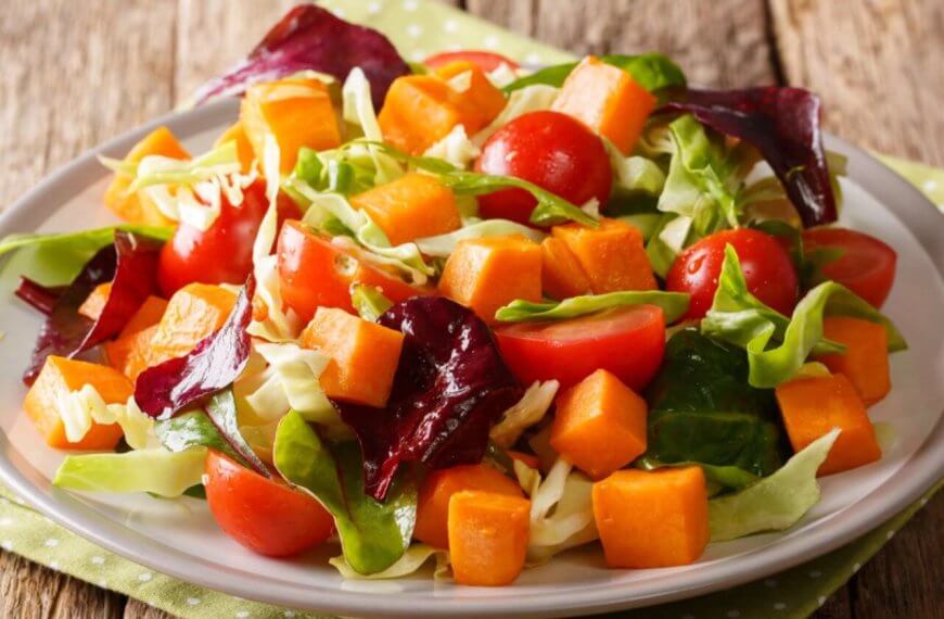 A fresh salad on a white plate features vibrant ingredients, including cherry tomatoes, cubed sweet potatoes, chopped cabbage, and a mix of green and purple leafy greens. Perfect for any food lover, this dish reflects the rich produce of Benguet Province. The plate rests on a green polka-dotted napkin on a rustic wooden table.