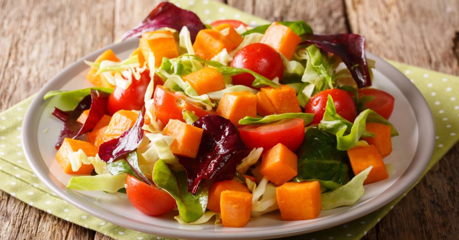A fresh salad on a white plate features vibrant ingredients, including cherry tomatoes, cubed sweet potatoes, chopped cabbage, and a mix of green and purple leafy greens. Perfect for any food lover, this dish reflects the rich produce of Benguet Province. The plate rests on a green polka-dotted napkin on a rustic wooden table.