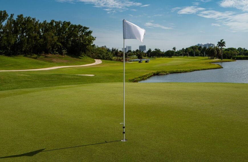 A golf course with a close-up view of a hole marked by a white flag on a green at one of the best golf and country clubs in the Philippines. In the background, there's a fairway surrounded by trees, a sand bunker, and a water area on the right. The sky is mostly clear with distant buildings visible beyond the trees.