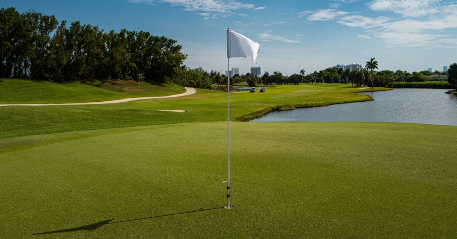 A golf course with a close-up view of a hole marked by a white flag on a green at one of the best golf and country clubs in the Philippines. In the background, there's a fairway surrounded by trees, a sand bunker, and a water area on the right. The sky is mostly clear with distant buildings visible beyond the trees.