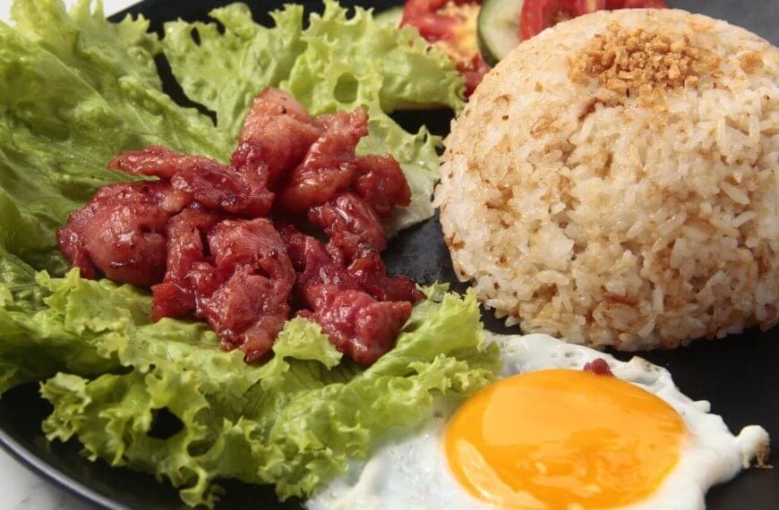 A black plate holds garlic fried rice topped with crushed garlic, accompanied by a sunny-side-up egg and pieces of caramelized pork placed over fresh green lettuce leaves. This classic Tapsilog, a popular Filipino breakfast in Metro Manila, is garnished with slices of tomato and cucumber in the background.