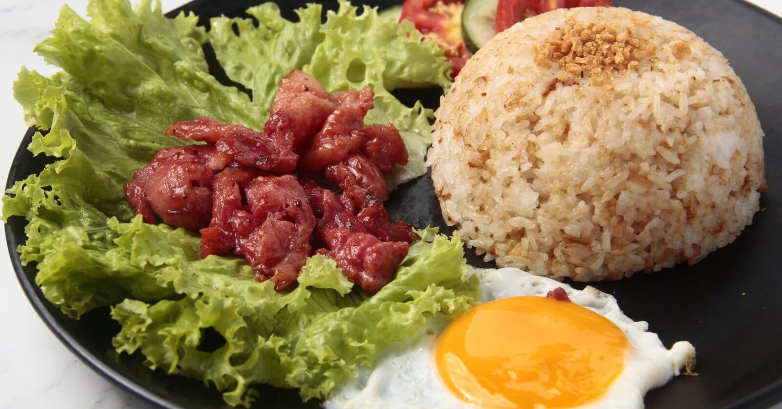 A black plate holds garlic fried rice topped with crushed garlic, accompanied by a sunny-side-up egg and pieces of caramelized pork placed over fresh green lettuce leaves. This classic Tapsilog, a popular Filipino breakfast in Metro Manila, is garnished with slices of tomato and cucumber in the background.