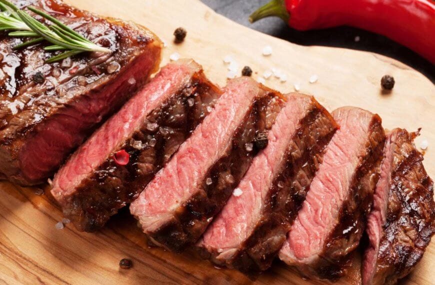 A cooked steak, partially sliced into pieces, is presented on a wooden cutting board, garnished with coarse sea salt and black peppercorns. A sprig of rosemary rests on the whole part of the steak. In the background at Bonifacio High Street's top restaurants in BGC, whole red and yellow chili peppers are visible.
