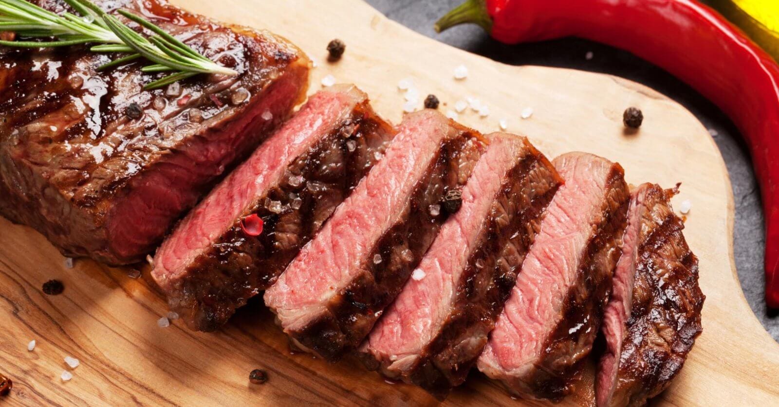 A cooked steak, partially sliced into pieces, is presented on a wooden cutting board, garnished with coarse sea salt and black peppercorns. A sprig of rosemary rests on the whole part of the steak. In the background at Bonifacio High Street's top restaurants in BGC, whole red and yellow chili peppers are visible.