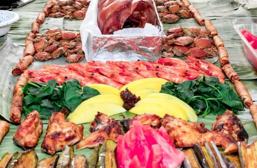 A festive spread of Filipino food is served on banana leaves at one of Metro Manila's top Boodle Fight Restaurants. Central to the layout are roasted pork, grilled chicken, shrimp, and vibrant fruits like mango and watermelon. Accompanying these are fresh greens, crabs, various grilled vegetables, and spring rolls arranged in an inviting manner.