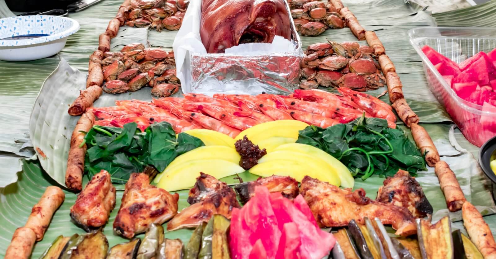 A festive spread of Filipino food is served on banana leaves at one of Metro Manila's top Boodle Fight Restaurants. Central to the layout are roasted pork, grilled chicken, shrimp, and vibrant fruits like mango and watermelon. Accompanying these are fresh greens, crabs, various grilled vegetables, and spring rolls arranged in an inviting manner.