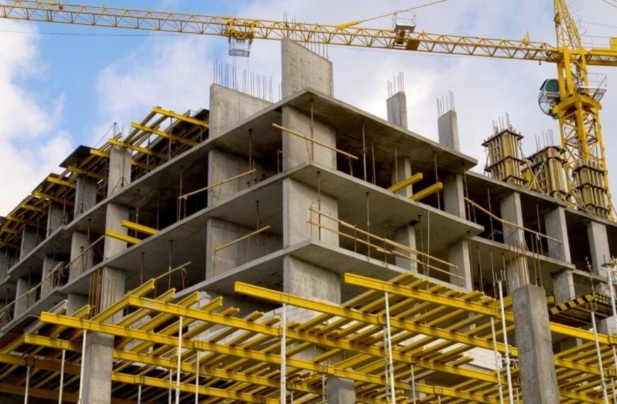 A construction site features a multi-story concrete building framework. Prominently visible are yellow scaffolding and support structures. Two tall tower cranes, essential for construction companies in the Philippines, are stationed nearby, one crane's arm extending over the building. The sky is partly cloudy with blue patches.