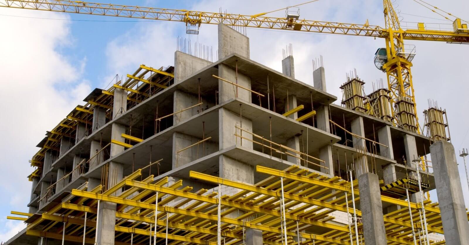 A construction site features a multi-story concrete building framework. Prominently visible are yellow scaffolding and support structures. Two tall tower cranes, essential for construction companies in the Philippines, are stationed nearby, one crane's arm extending over the building. The sky is partly cloudy with blue patches.