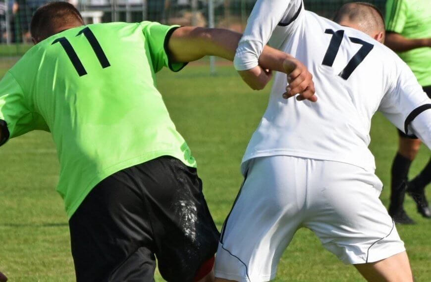 Two soccer players, each showcasing custom uniforms—one in a green jersey with the number 11 and the other in a white jersey with the number 17—compete for the ball on a grassy field. Both are leaning in and making contact with their arms as they balance on one leg, underlining their suppliers' quality craftsmanship. A goal post and fencing are visible in the background.