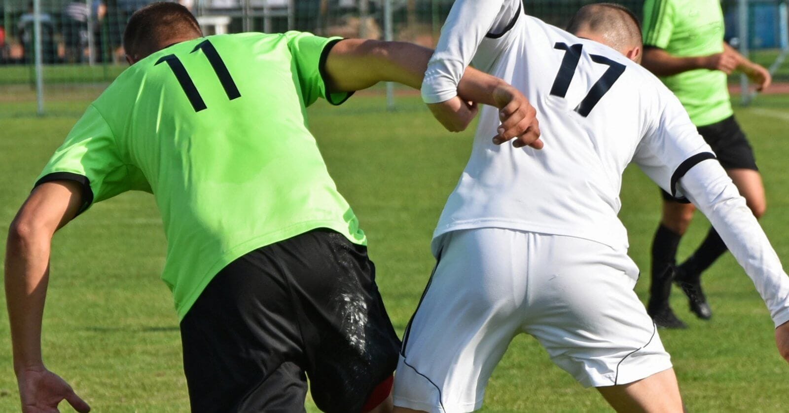Two soccer players, each showcasing custom uniforms—one in a green jersey with the number 11 and the other in a white jersey with the number 17—compete for the ball on a grassy field. Both are leaning in and making contact with their arms as they balance on one leg, underlining their suppliers' quality craftsmanship. A goal post and fencing are visible in the background.