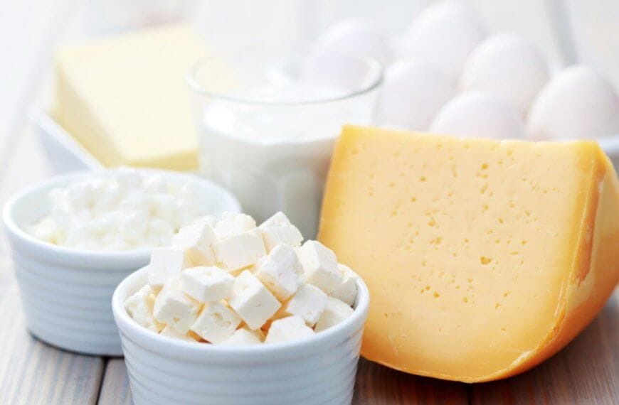 A wooden table in Metro Manila showcases a variety of dairy products supplied by local Dairy Product Suppliers, including a block of cheddar cheese, a bowl of white feta cheese cubes, cottage cheese, glass of milk, dish of butter, and a bowl of eggs. The items are elegantly arranged in white bowls and on plates.