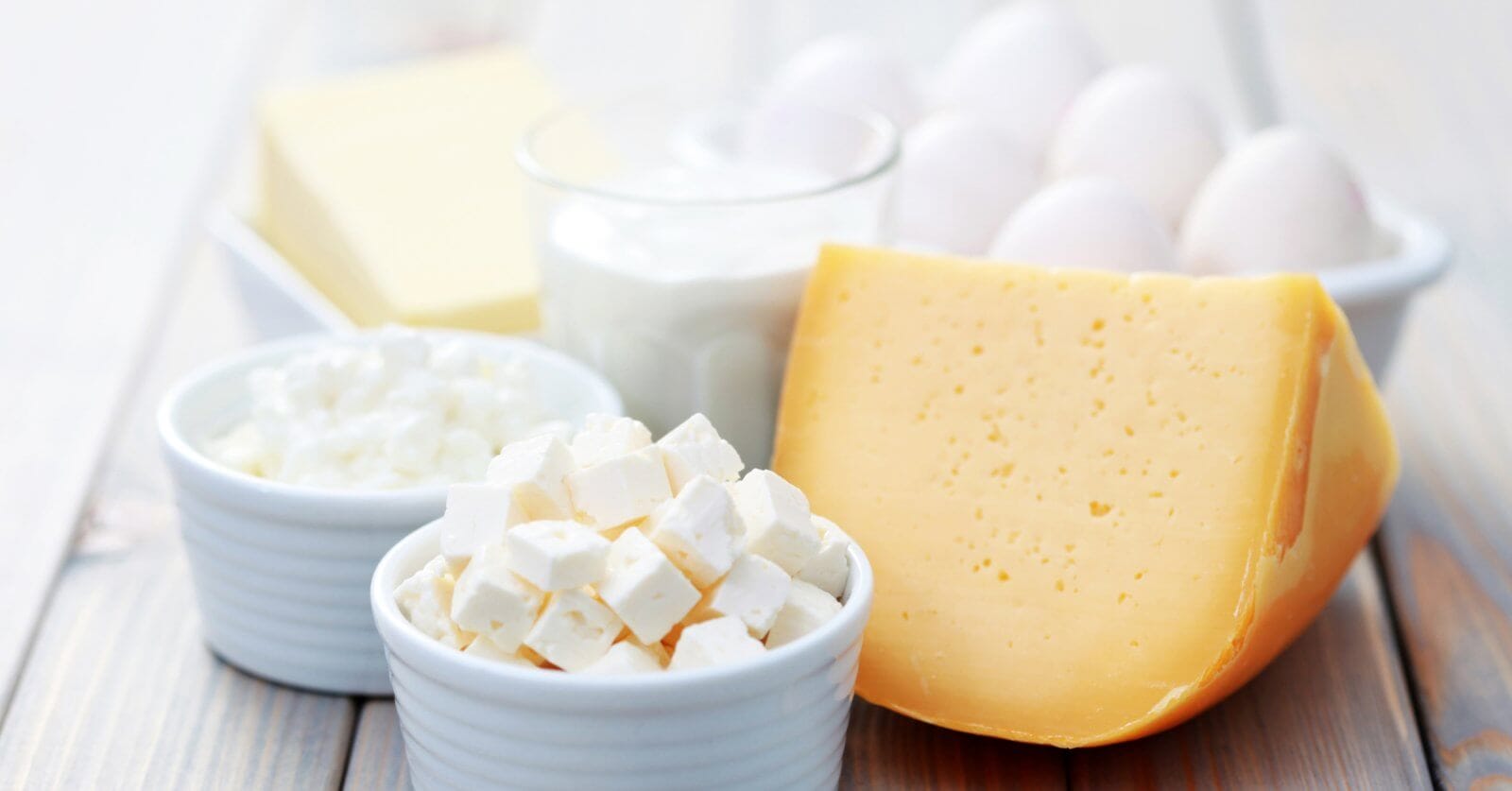 A wooden table in Metro Manila showcases a variety of dairy products supplied by local Dairy Product Suppliers, including a block of cheddar cheese, a bowl of white feta cheese cubes, cottage cheese, glass of milk, dish of butter, and a bowl of eggs. The items are elegantly arranged in white bowls and on plates.