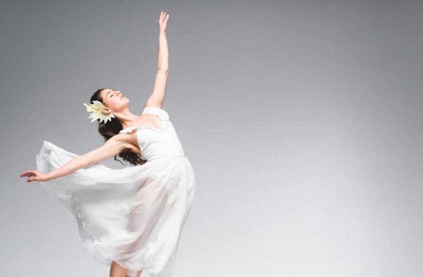 A ballet dancer in a flowing white dress and flower headpiece gracefully poses against a plain grey background. Her dress billows around her as she stands en pointe, creating a sense of motion and elegance—a true representation of the best dance studios in Metro Manila.