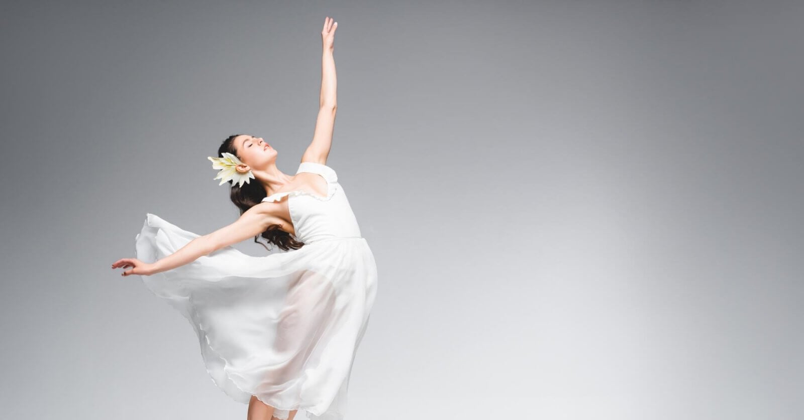 A ballet dancer in a flowing white dress and flower headpiece gracefully poses against a plain grey background. Her dress billows around her as she stands en pointe, creating a sense of motion and elegance—a true representation of the best dance studios in Metro Manila.