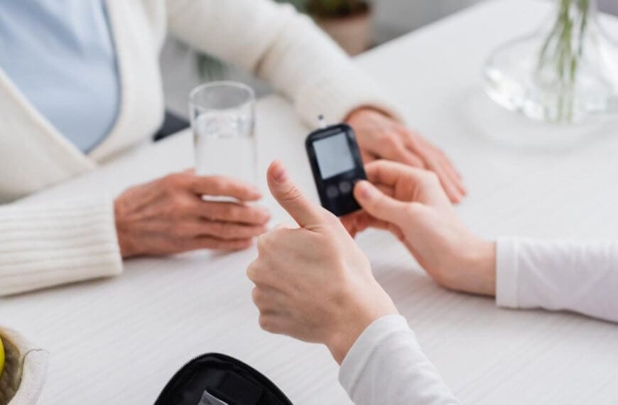 Two people sit at a white table in a bustling Metro Manila clinic. One person holds a blood glucose meter, giving a thumbs-up, while the other holds a glass of water. On the table are a zippered case with medical supplies and a bowl with green apples. The background is blurred, focusing on their interaction.