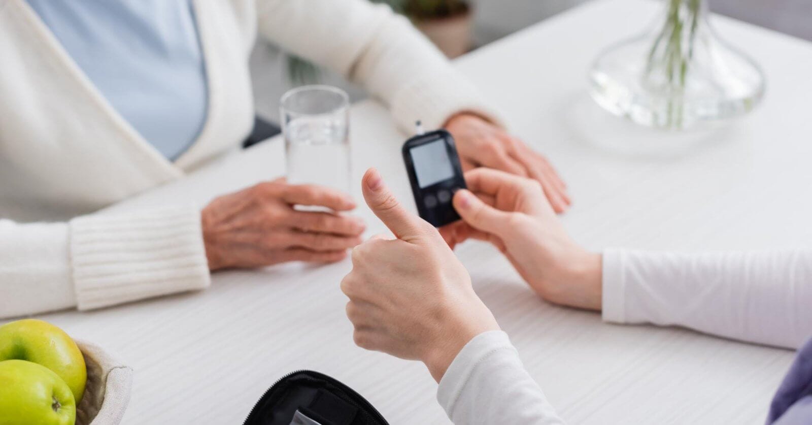 Two people sit at a white table in a bustling Metro Manila clinic. One person holds a blood glucose meter, giving a thumbs-up, while the other holds a glass of water. On the table are a zippered case with medical supplies and a bowl with green apples. The background is blurred, focusing on their interaction.
