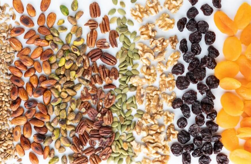 A top-view photo displays an assortment of nuts, seeds, and dried fruits arranged in vertical rows. From left to right: sunflower seeds, almonds, pistachios, pecans, pumpkin seeds, walnuts, dried cranberries, and dried apricots—all sourced from top nut suppliers in Metro Manila—set against a white background.